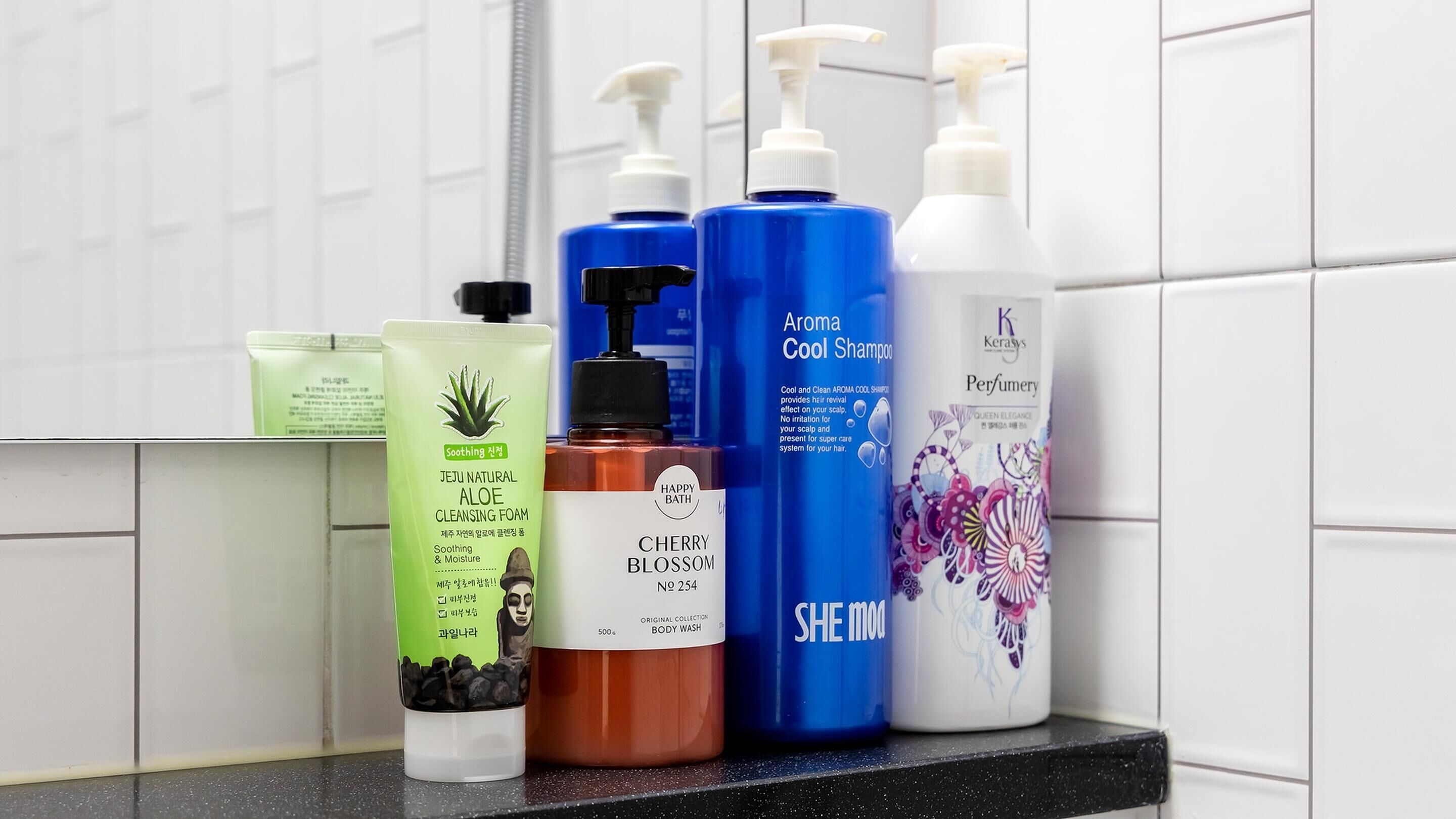 Kylpyhuoneen hyllyllä on erilaisia hygieniatuotteita, kuten vihreää aloe veraa sisältävää puhdistusvaahtoa, sinistä suihkugeeliä ja valkoinen hajuvesipullo.
