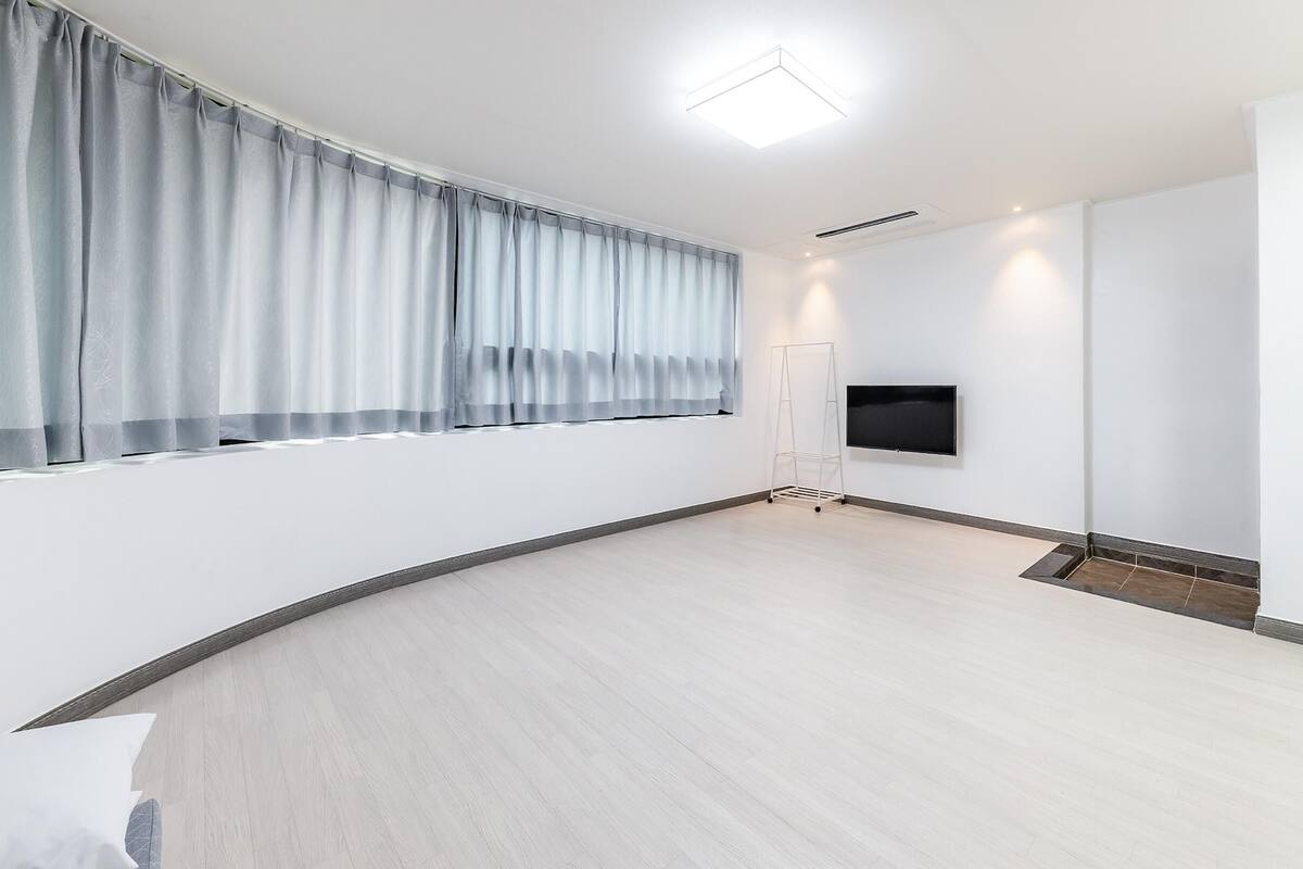 A spacious room with a large window, a flat-screen TV mounted on the wall, and a light-colored wooden floor.