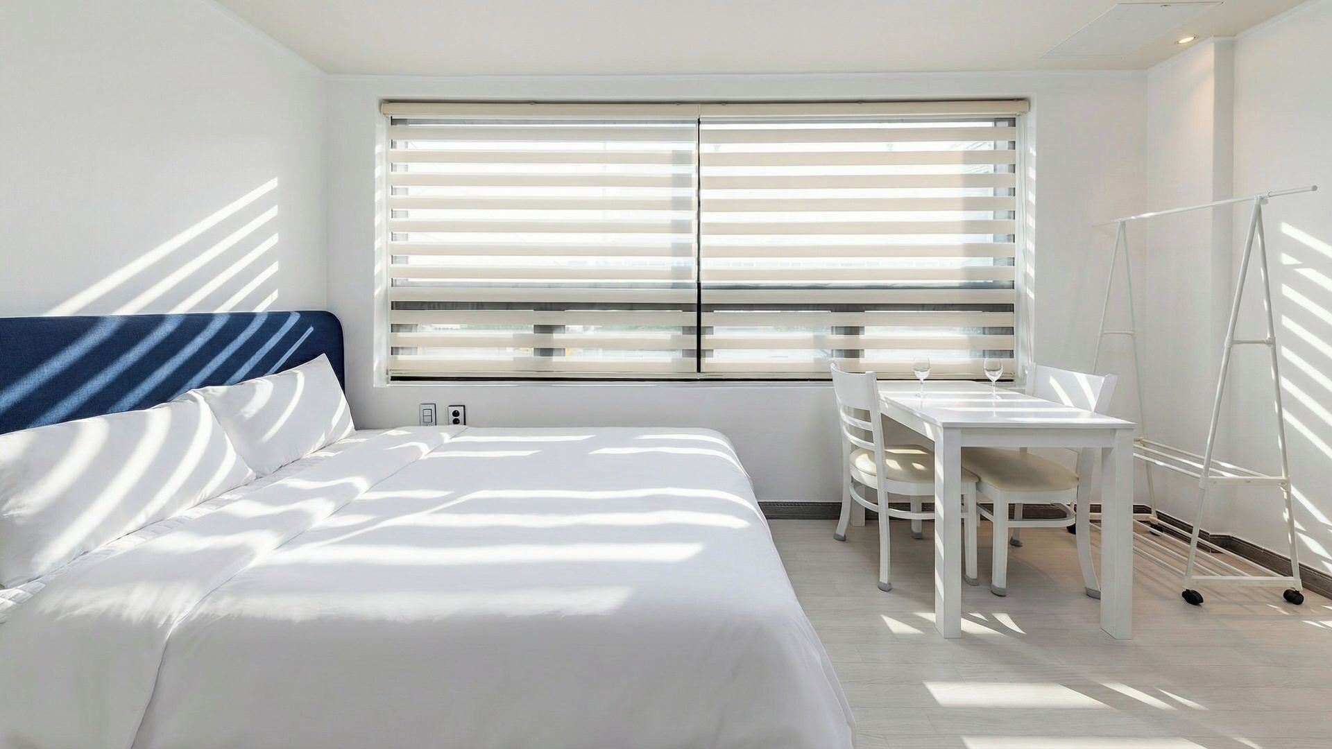 A hotel room with a bed, a desk, and a chair. There is a window with blinds, and a ladder is leaning against the wall.