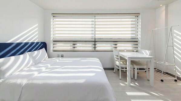 A hotel room with a bed, a desk, and a chair. There is a window with blinds, and a ladder is leaning against the wall.