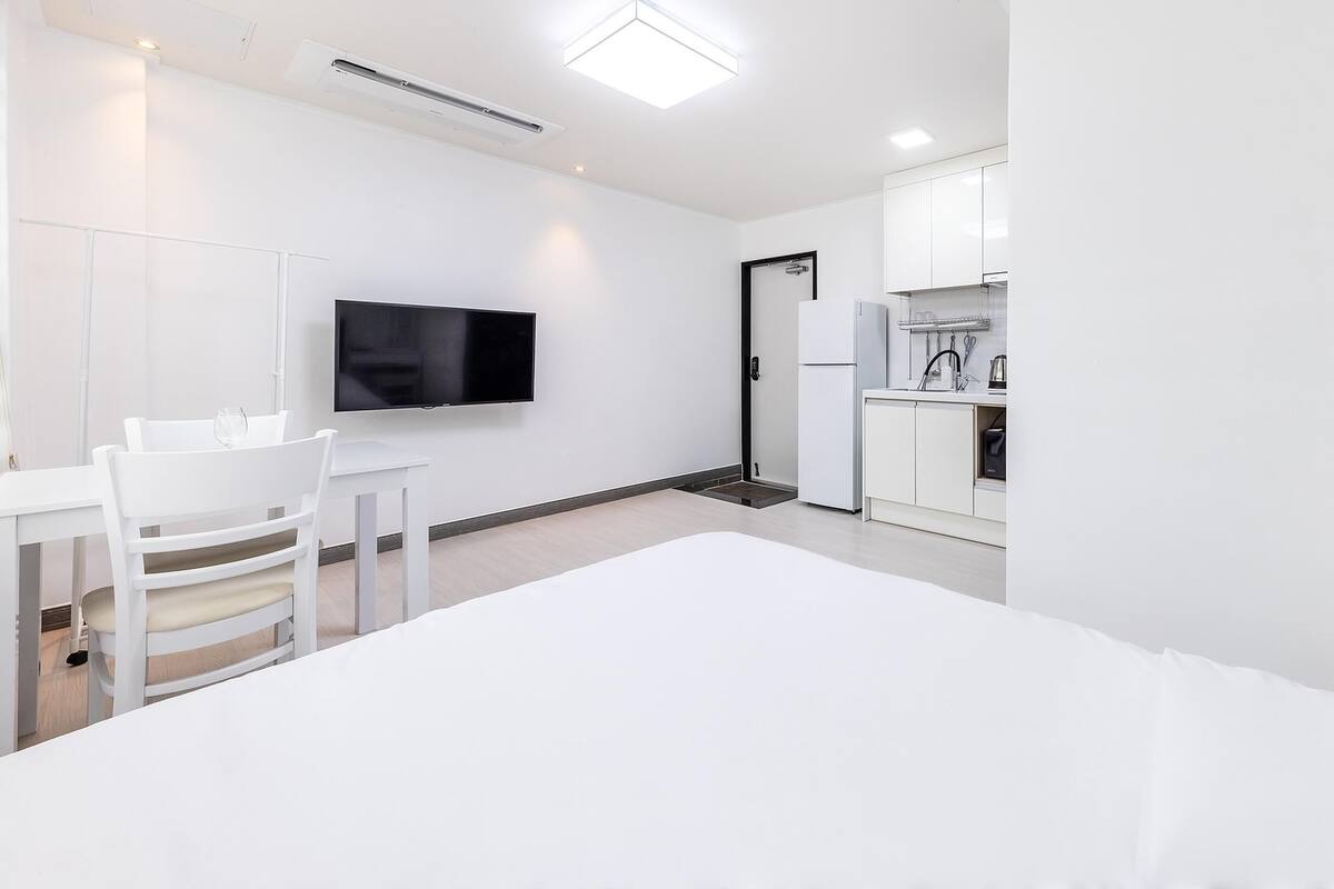 A modern hotel room with a bed, a desk with a chair, a television, a kitchen area with a refrigerator and a sink, and a door leading to the bathroom.