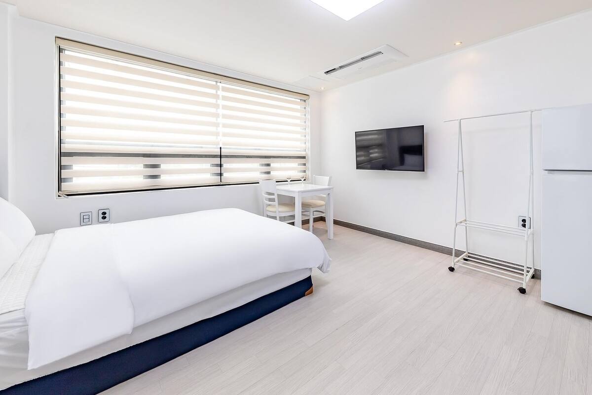 A modern hotel room with a bed, a desk with a chair, a TV mounted on the wall, and a wardrobe.