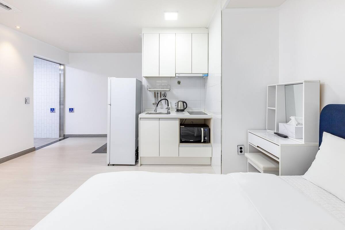 A modern hotel room with a bed, a small kitchenette with a microwave, a refrigerator, and a bedside table with a lamp.