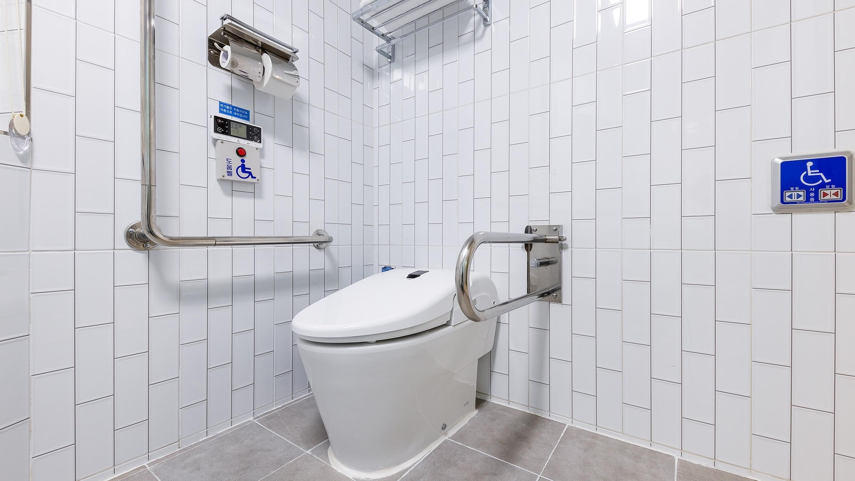 A white tiled bathroom with a white toilet, a handrail, and a sign indicating accessibility.
