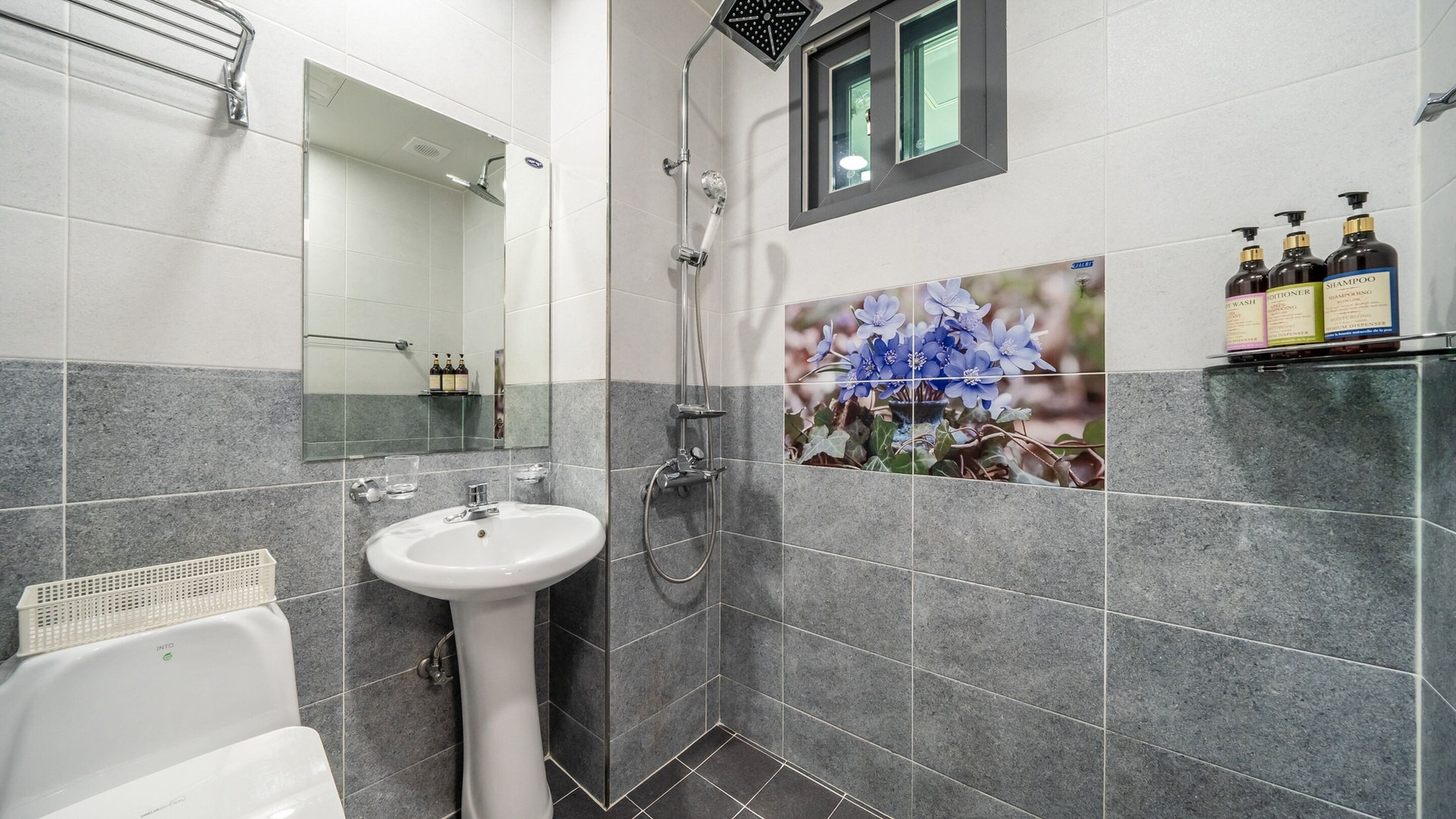 Un baño moderno con un lavamanos blanco, ducha y espejo. Hay artículos de aseo en la pared y una ventana sobre el espejo.