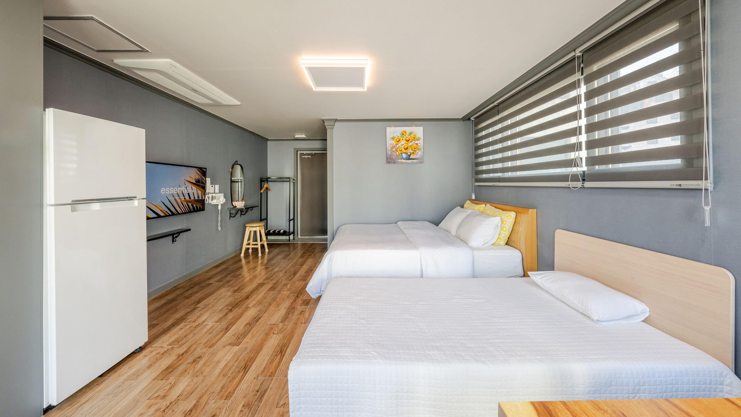A compact hotel room with two beds, a refrigerator, a small stool, a bench, a wall-mounted TV, and a window with blinds.