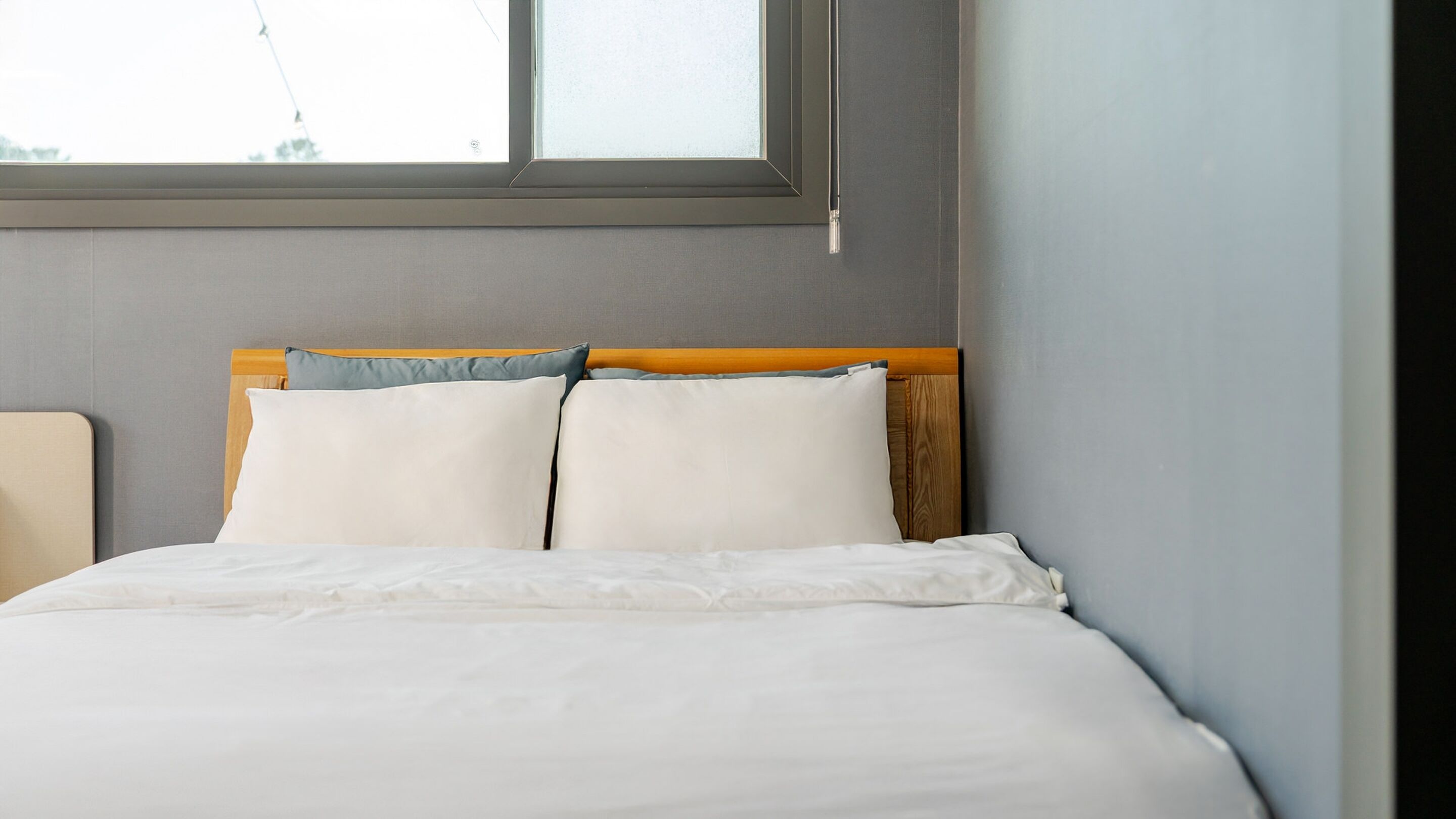 Una cama bien hecha con ropa blanca y un cabecero de madera, una ventana encima y una pared gris.