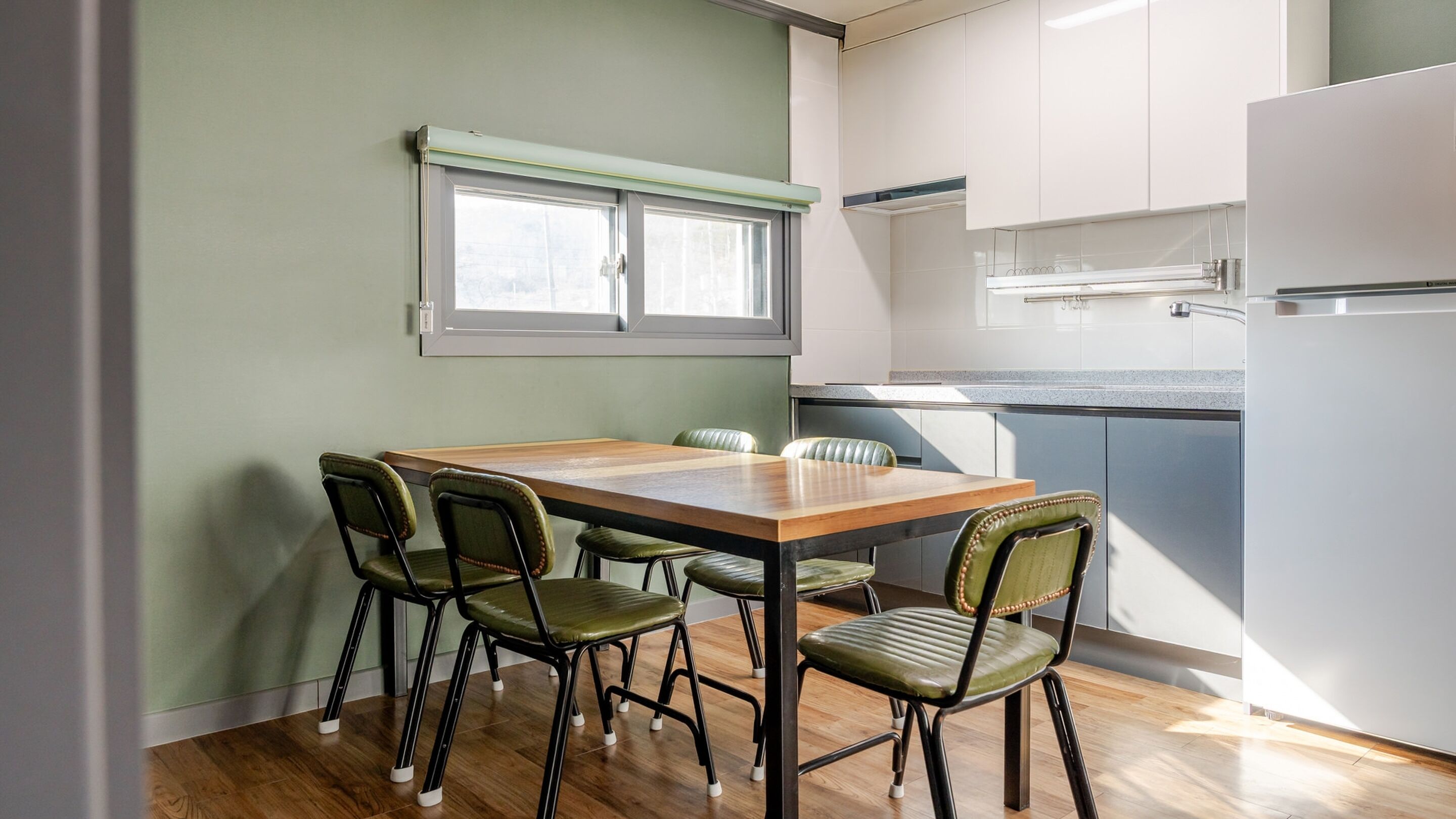 Uma sala de jantar com mesa de madeira e quatro cadeiras verdes, janela com persianas, e área de cozinha com armários brancos e uma geladeira branca.