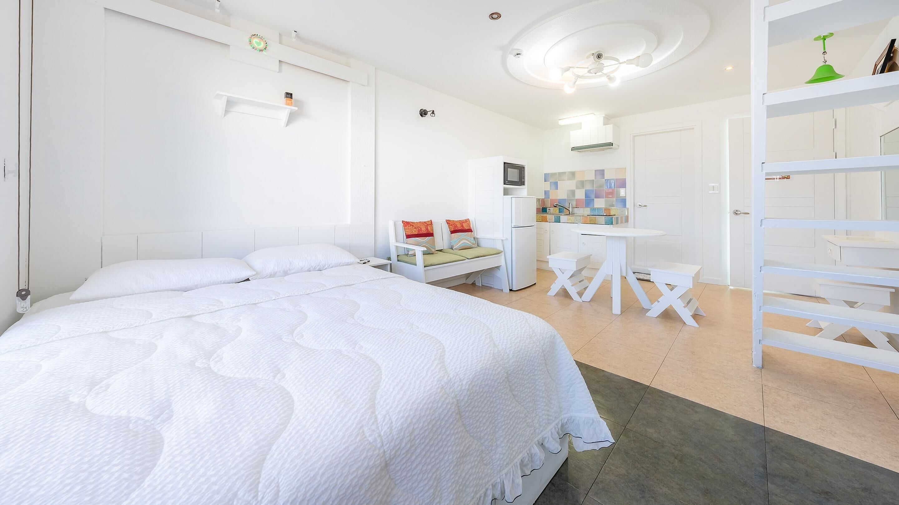 A modern, white-themed bedroom with a large bed, a small desk, and a kitchenette area.