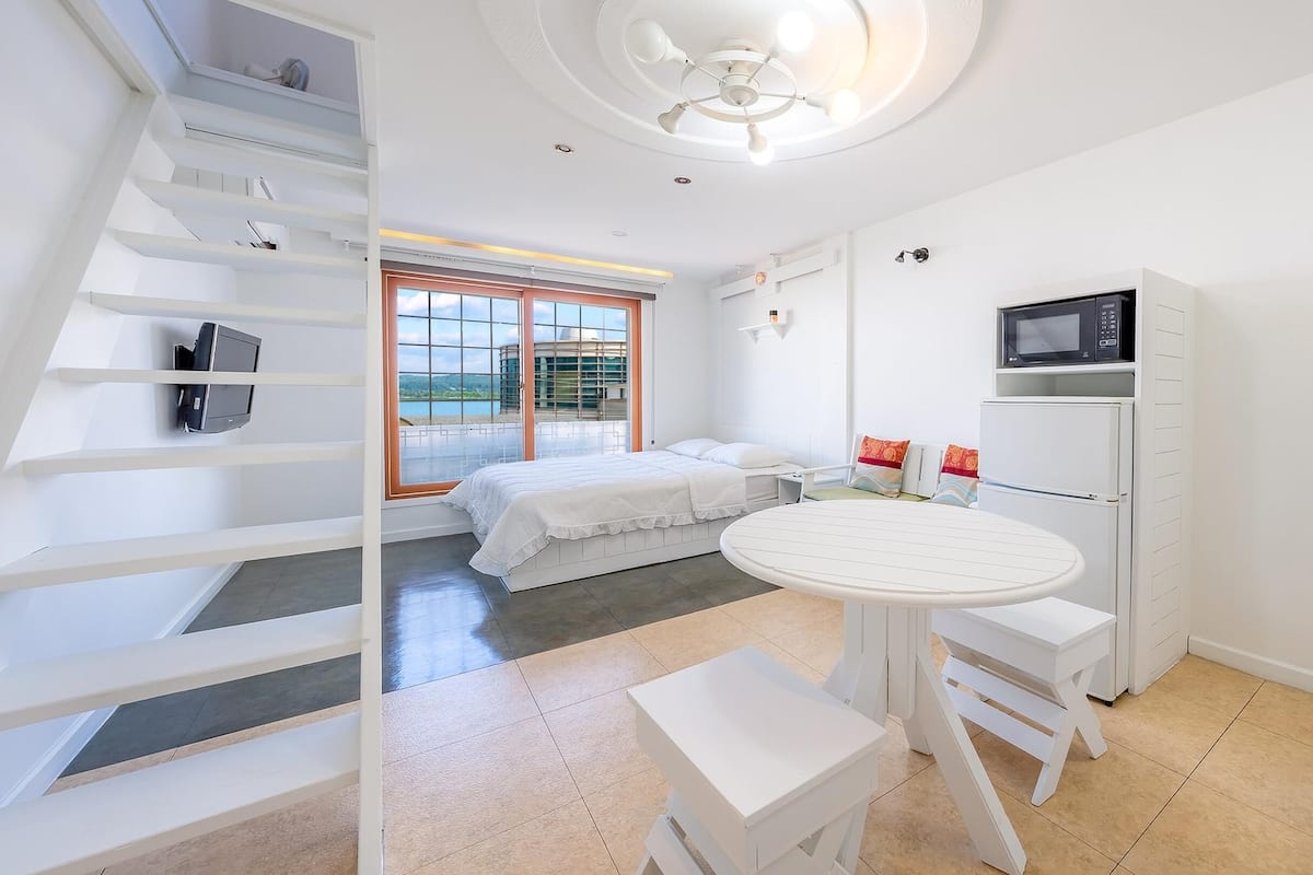 Une chambre à coucher moderne, aux tons blancs, comprenant un lit, une petite table et une chaise. Une fenêtre offre une vue sur l’extérieur.