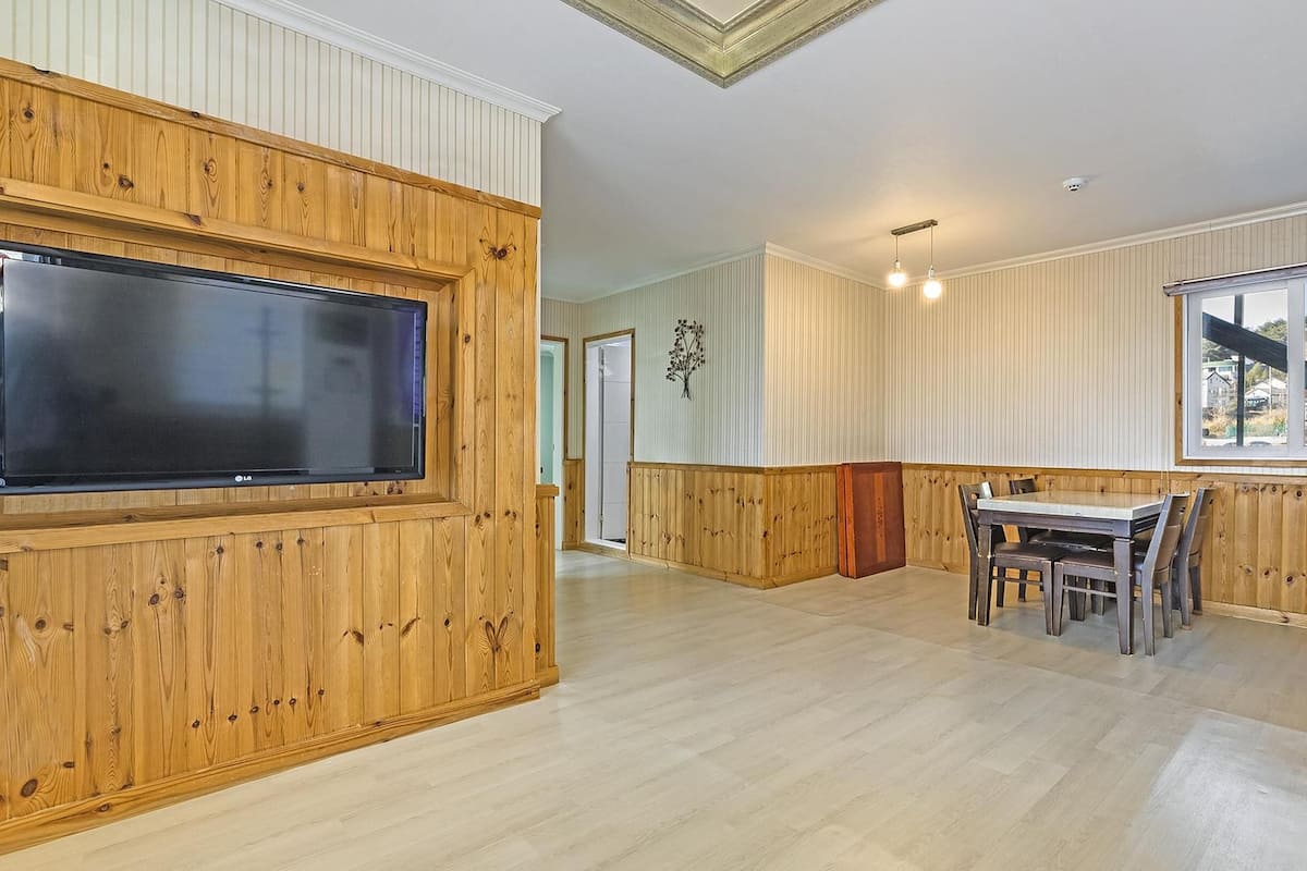 Un vaste salon doté d’un grand téléviseur à écran plat fixé à un mur en bois, d’un coin repas avec une table et des chaises, et de grandes fenêtres offrant une vue sur l’extérieur.