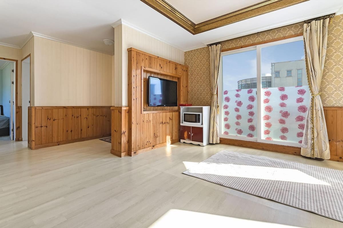 Une pièce spacieuse avec des lambris en bois, un téléviseur à écran plat fixé au mur, un four à micro-ondes et une grande fenêtre agrémentée de rideaux à motifs.