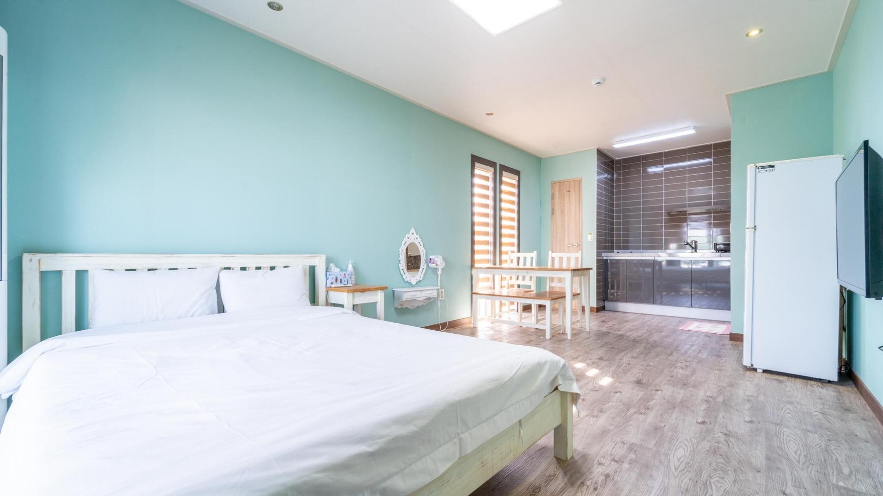 A bedroom with a bed, a small table with chairs, a mirror, and a kitchenette with a refrigerator and stove.