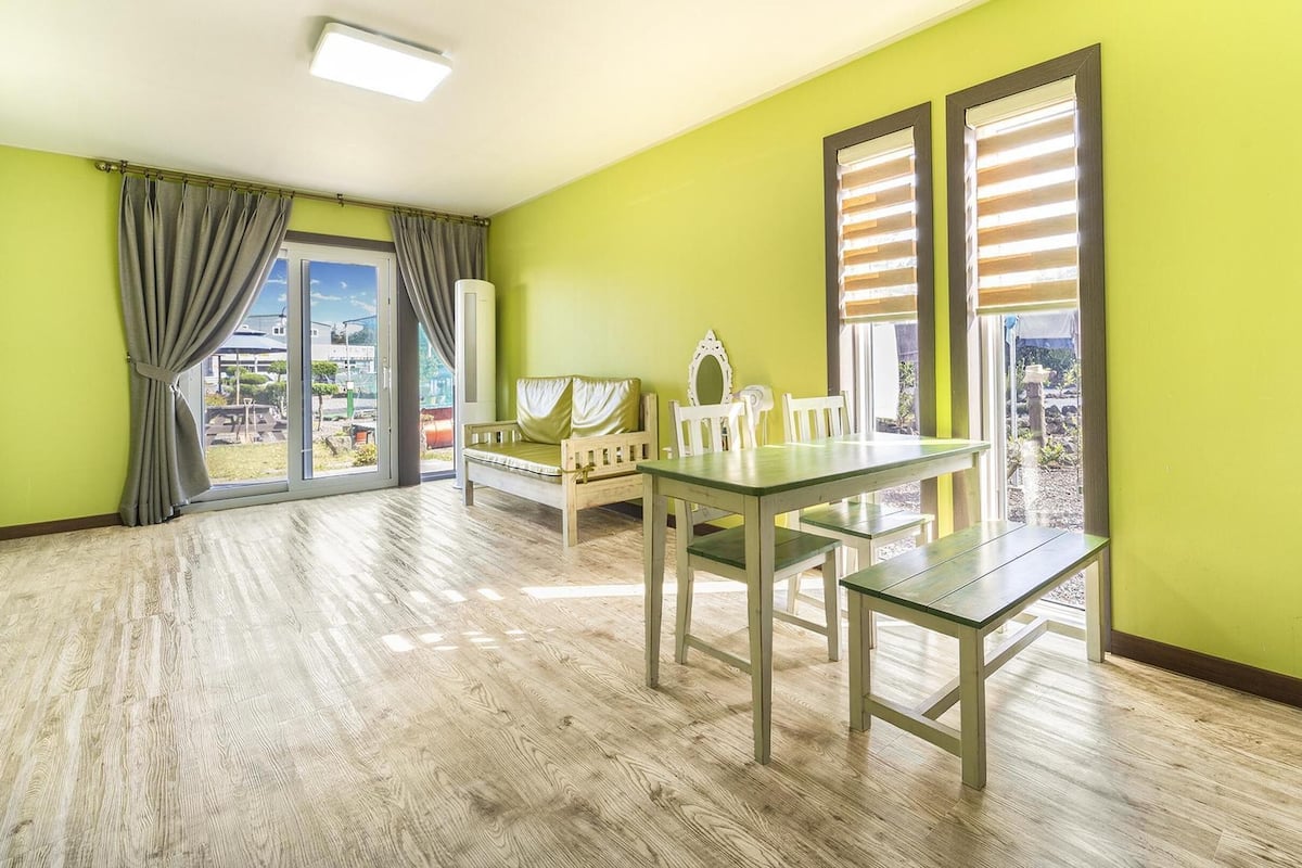 A bright room with a wooden floor, a green wall, a table with benches, and a window with blinds.