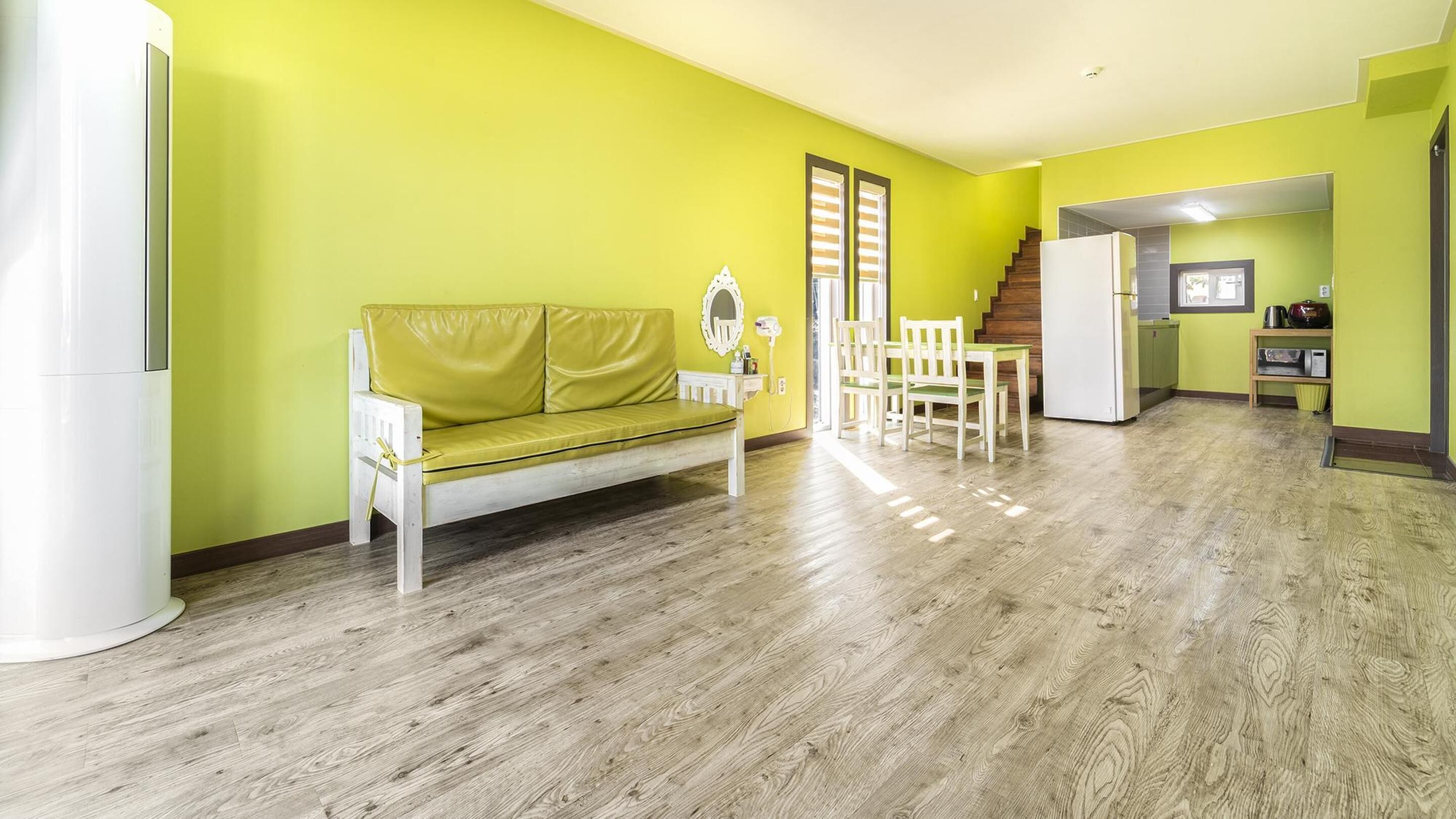 Een lichte gele kamer met een witte bank, houten vloeren en een keuken met een koelkast en fornuis.