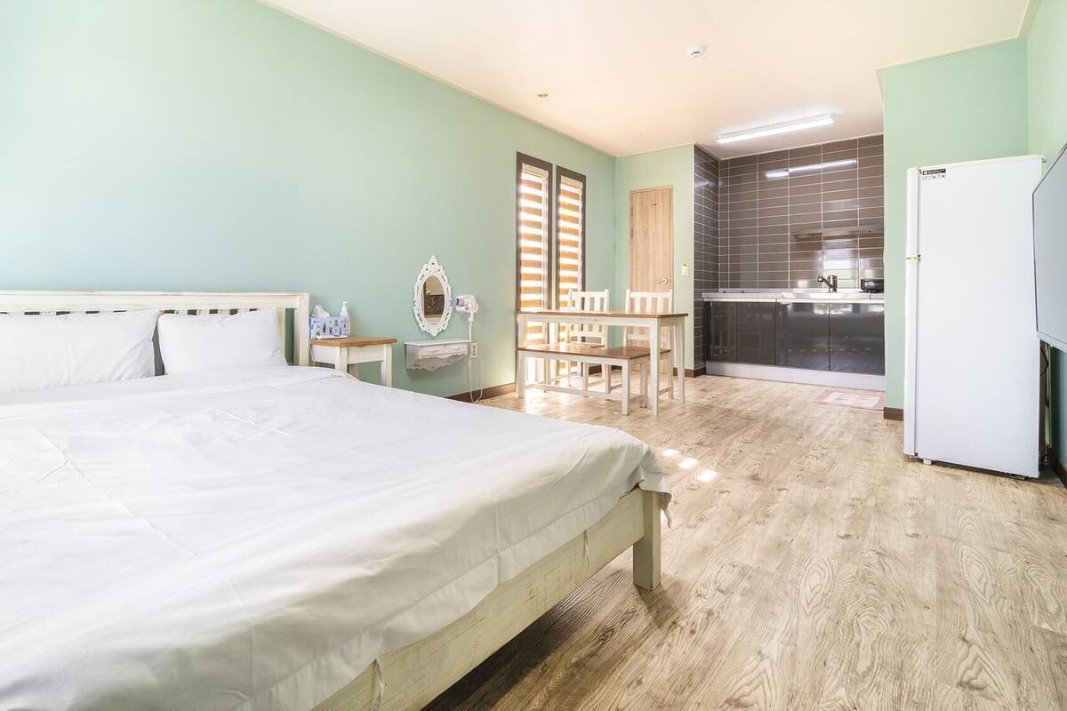 A bedroom with a bed, a desk, a chair, a mirror, a built-in kitchen with a refrigerator, and a TV.