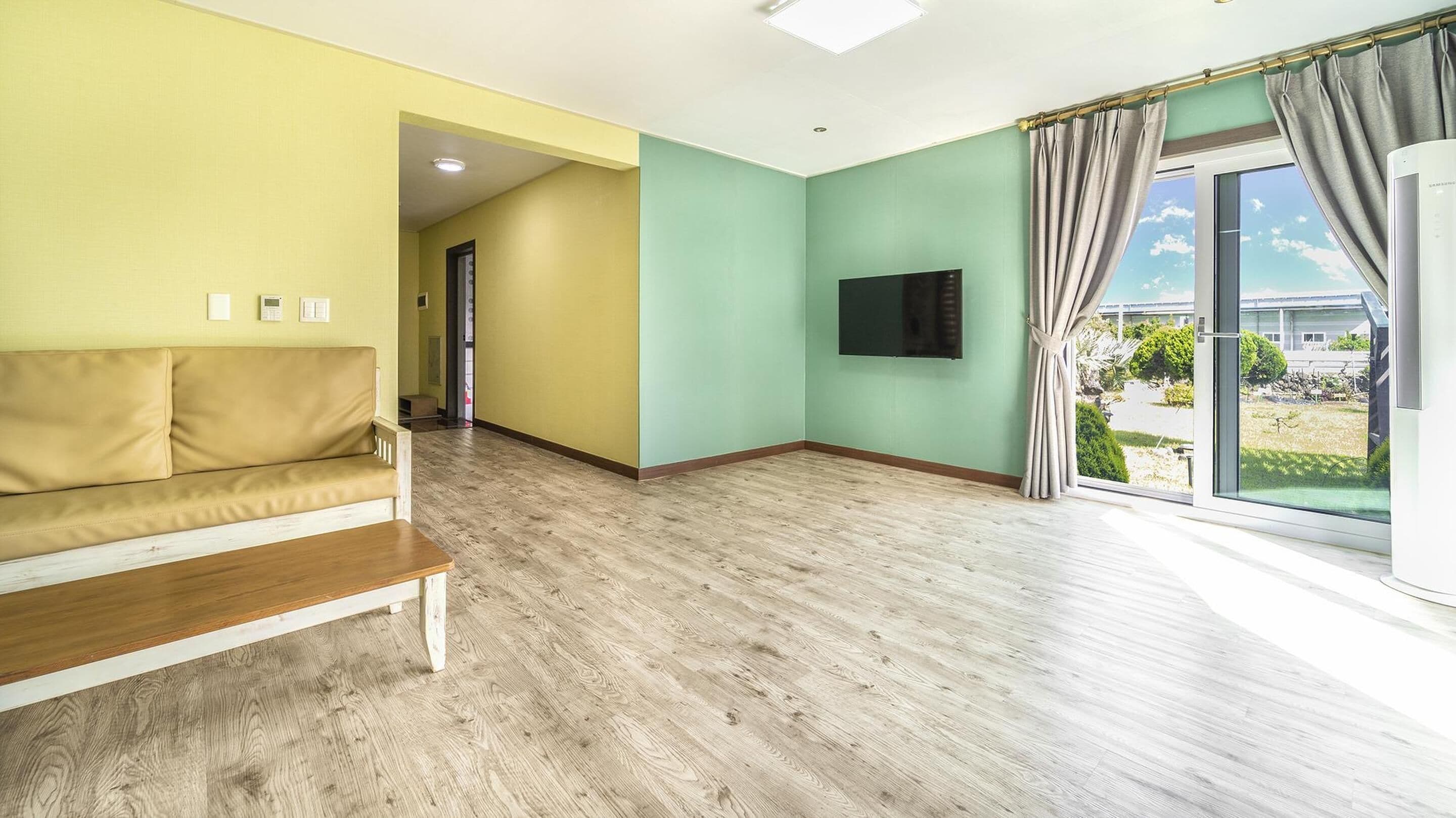 Ein Wohnzimmer mit beiger Couch, einem Holztisch, einem an der Wand befestigten Flachbildfernseher und einem großen Fenster mit Vorhängen.