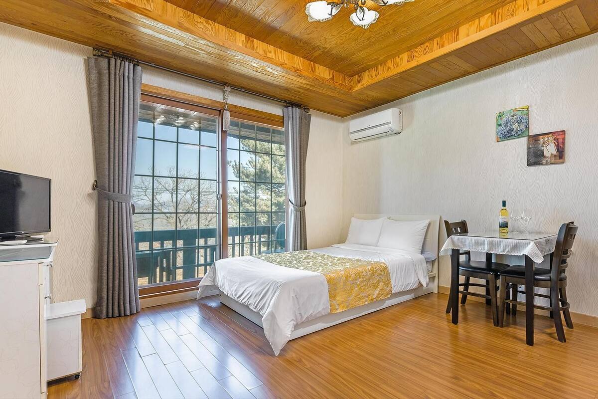A bedroom with a bed, a desk with a chair, a television, and an air conditioning unit.