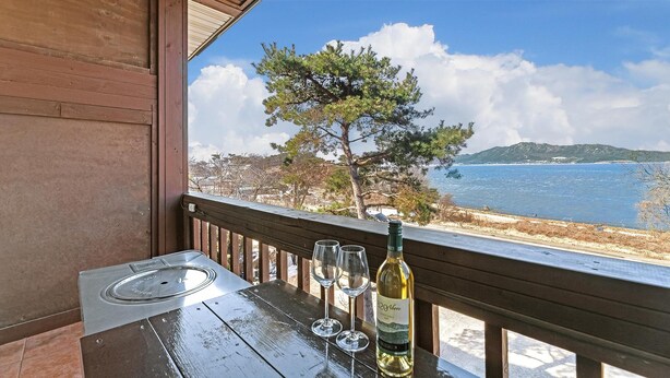 A wooden table with a bottle of wine and two glasses on a balcony overlooking a coastal view.
