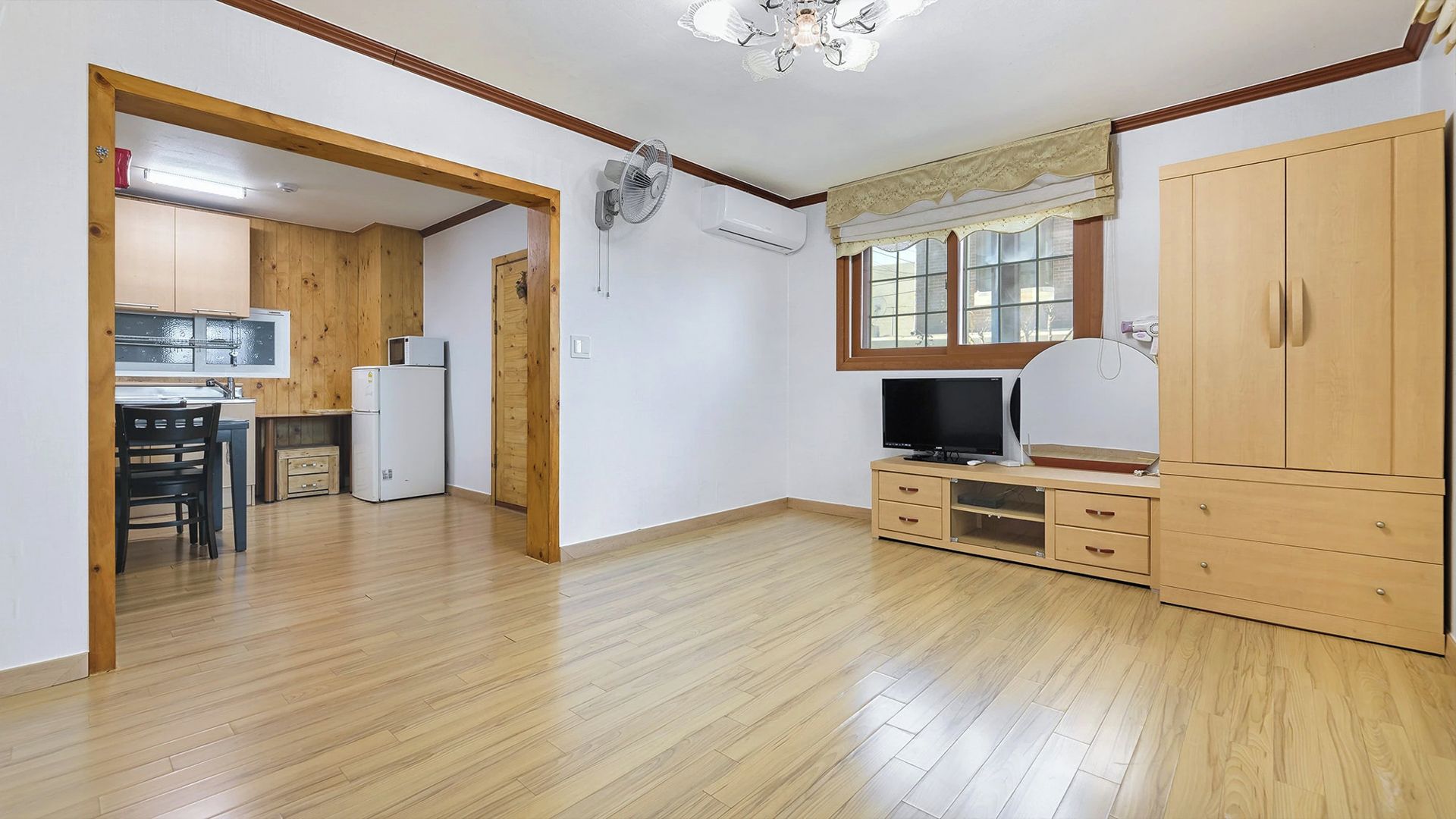 Une pièce avec un plancher en bois, un téléviseur posé sur un meuble en bois, un coin cuisine avec des placards et un réfrigérateur, et une fenêtre avec des rideaux.