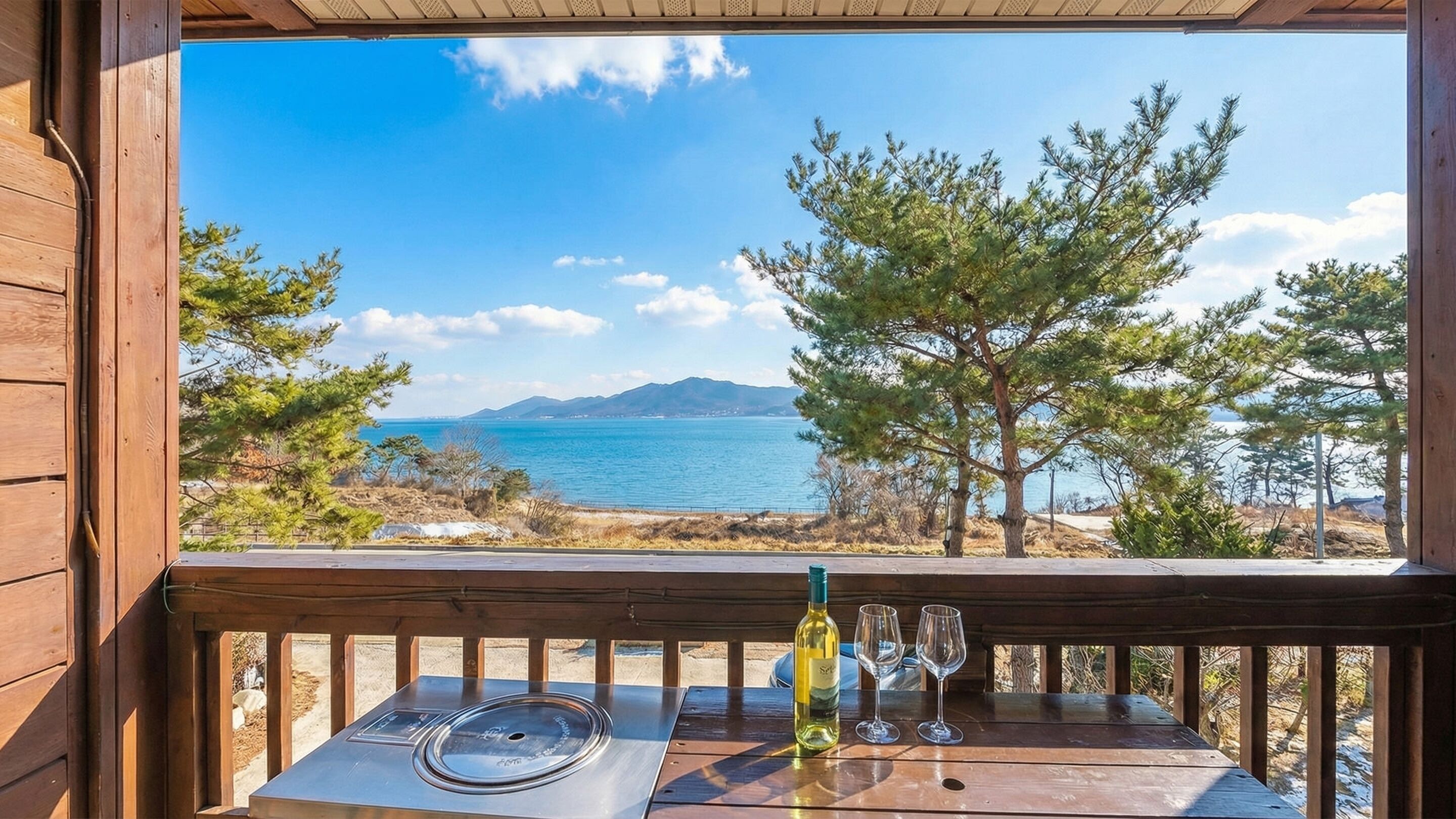 A wooden deck with a table, a bottle of wine, and two glasses. The deck overlooks a coastal landscape with trees and a view of the sea.