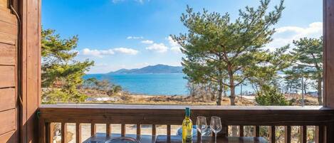 A wooden deck with a table, a bottle of wine, and two glasses. The deck overlooks a coastal landscape with trees and a view of the sea.