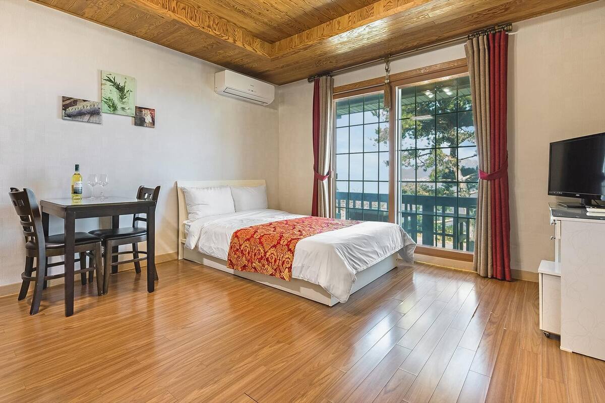 A hotel room with a wooden floor, a bed with a red and white bedspread, a desk with a chair, a television, and a window with curtains.