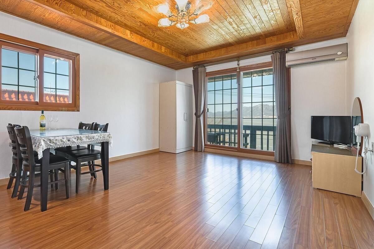 A spacious room with a wooden ceiling, a dining table with chairs, a TV, and a balcony with a view.