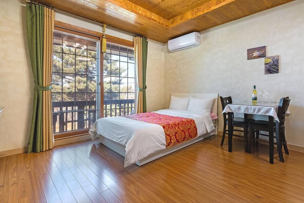 A bedroom with a bed, a table with two chairs, an air conditioning unit, and a window with curtains.