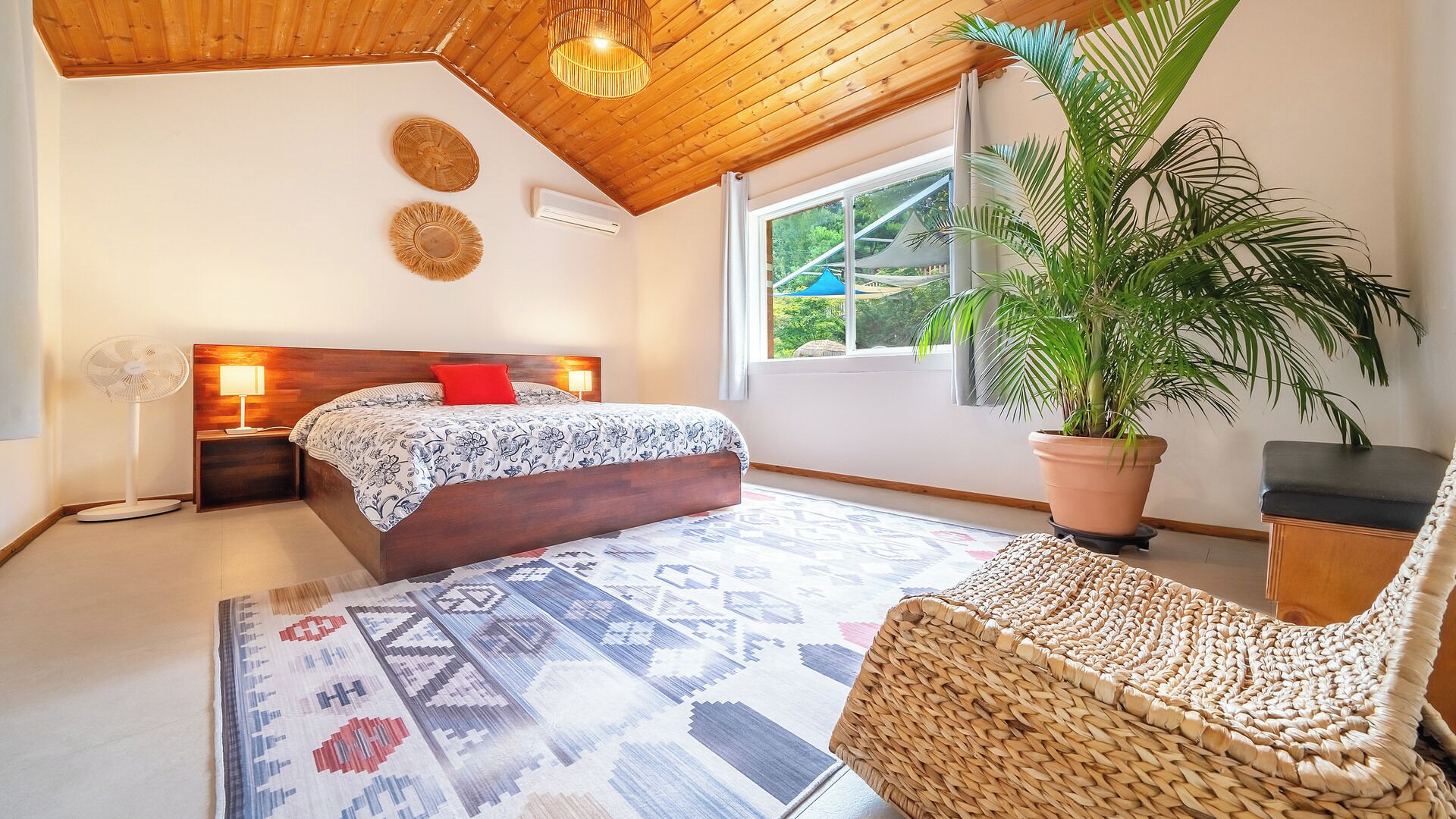 Un dormitorio con una cama, lámparas de noche, un sillón de mimbre, una planta en maceta y vistas al exterior con vegetación.