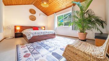 Un dormitorio con una cama, lámparas de noche, un sillón de mimbre, una planta en maceta y vistas al exterior con vegetación.