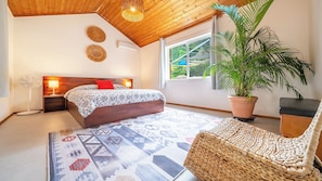 Un dormitorio con una cama, lámparas de noche, un sillón de mimbre, una planta en maceta y vistas al exterior con vegetación.