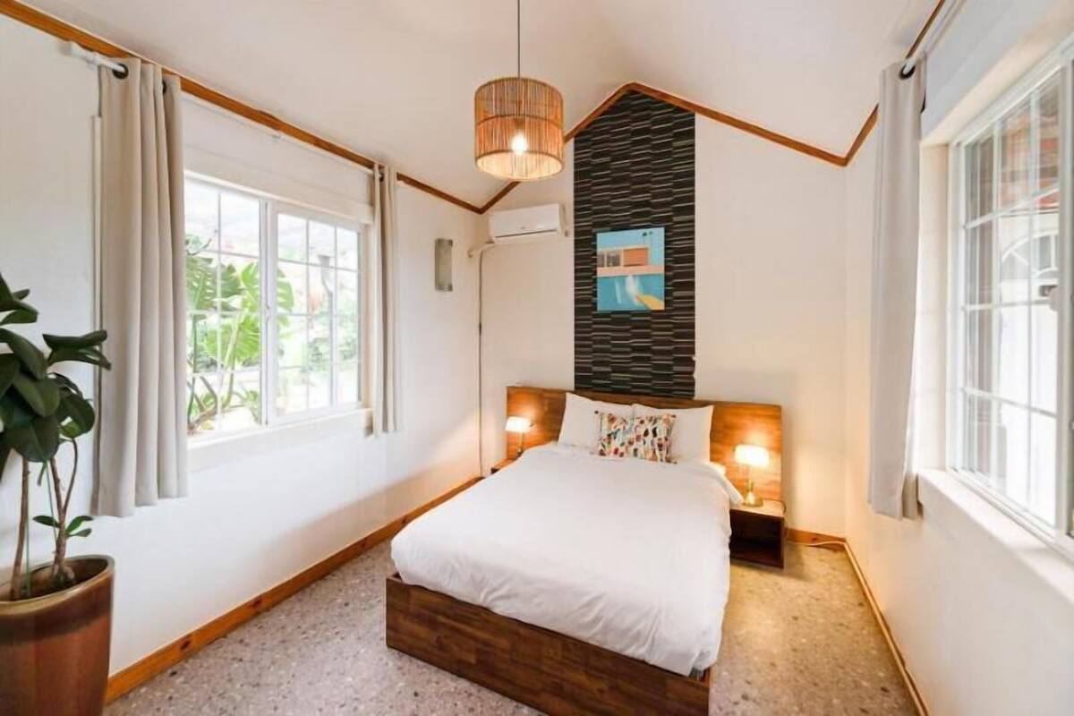 A bedroom with a bed, a potted plant, a wall-mounted air conditioner, and a window with curtains.