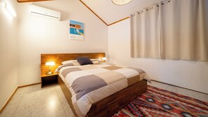 A bedroom with a wooden headboard, a bed with a checkered bedspread, a nightstand with a lamp, a wall-mounted air conditioner, and a window with curtains.
