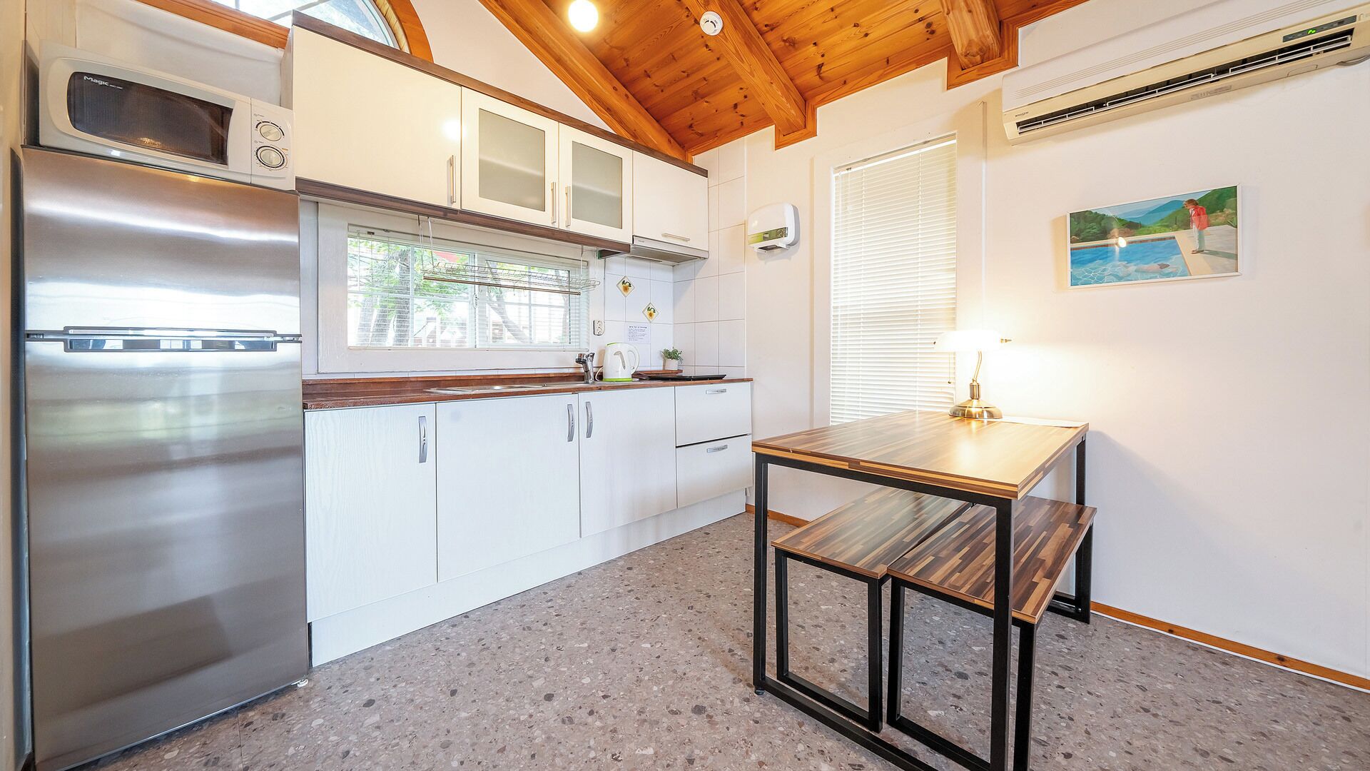 A kitchen with a stainless steel refrigerator, white cabinets, a wooden ceiling, and a small table with a lamp.