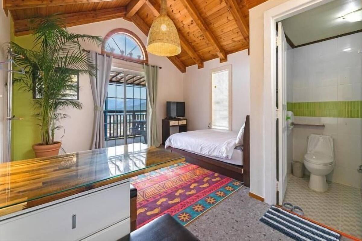 A bedroom with a bed, a desk, a potted plant, and a balcony.
