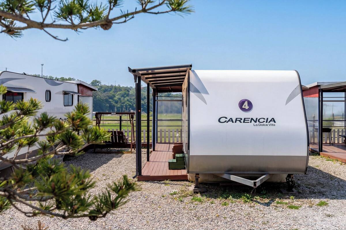 A caravan with a covered outdoor seating area, a white trailer with 'CARENCIA La Dolce Vita' branding, and a natural landscape in the background.