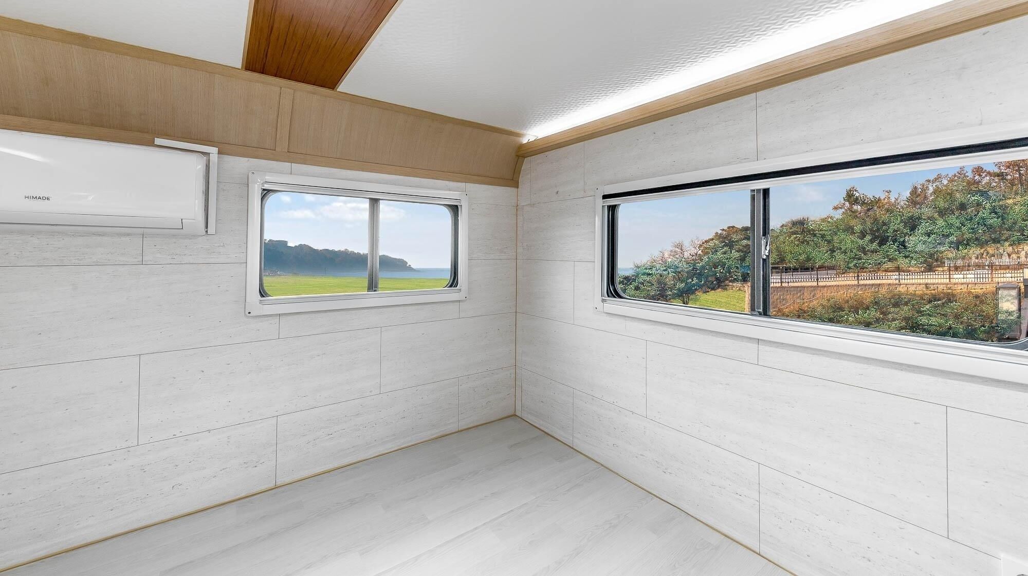 A room with white walls, a wooden ceiling, and two windows offering views of greenery and a distant landscape.