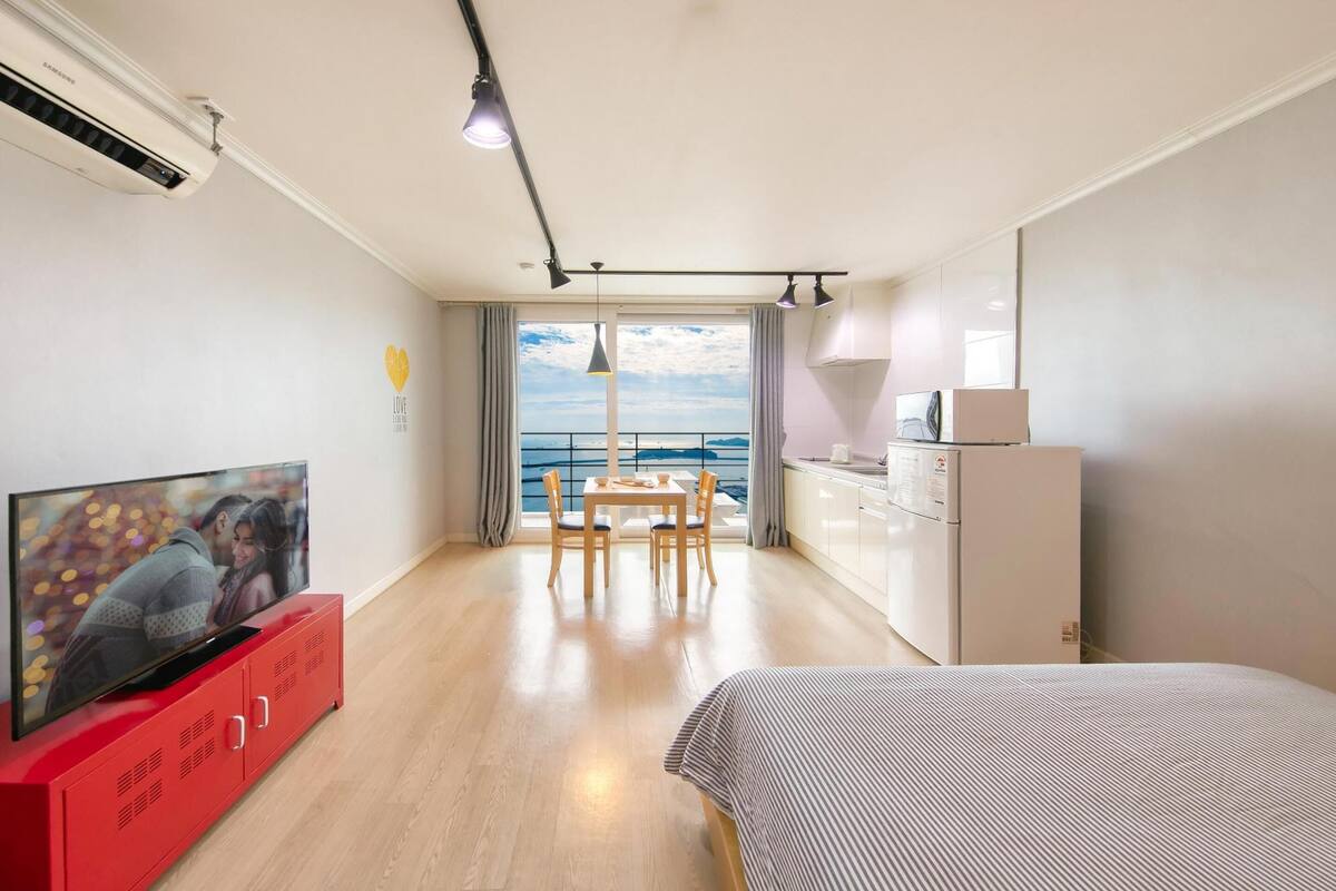 A modern hotel room with a TV on a red cabinet, a dining area with a table and chairs, a bed, a white wardrobe, and a view of the sea through large windows.