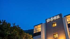 A modern building with illuminated signage, large windows, and a view of trees and hills in the background. - Yeosu Harushimpyo Pension (Yeosu)