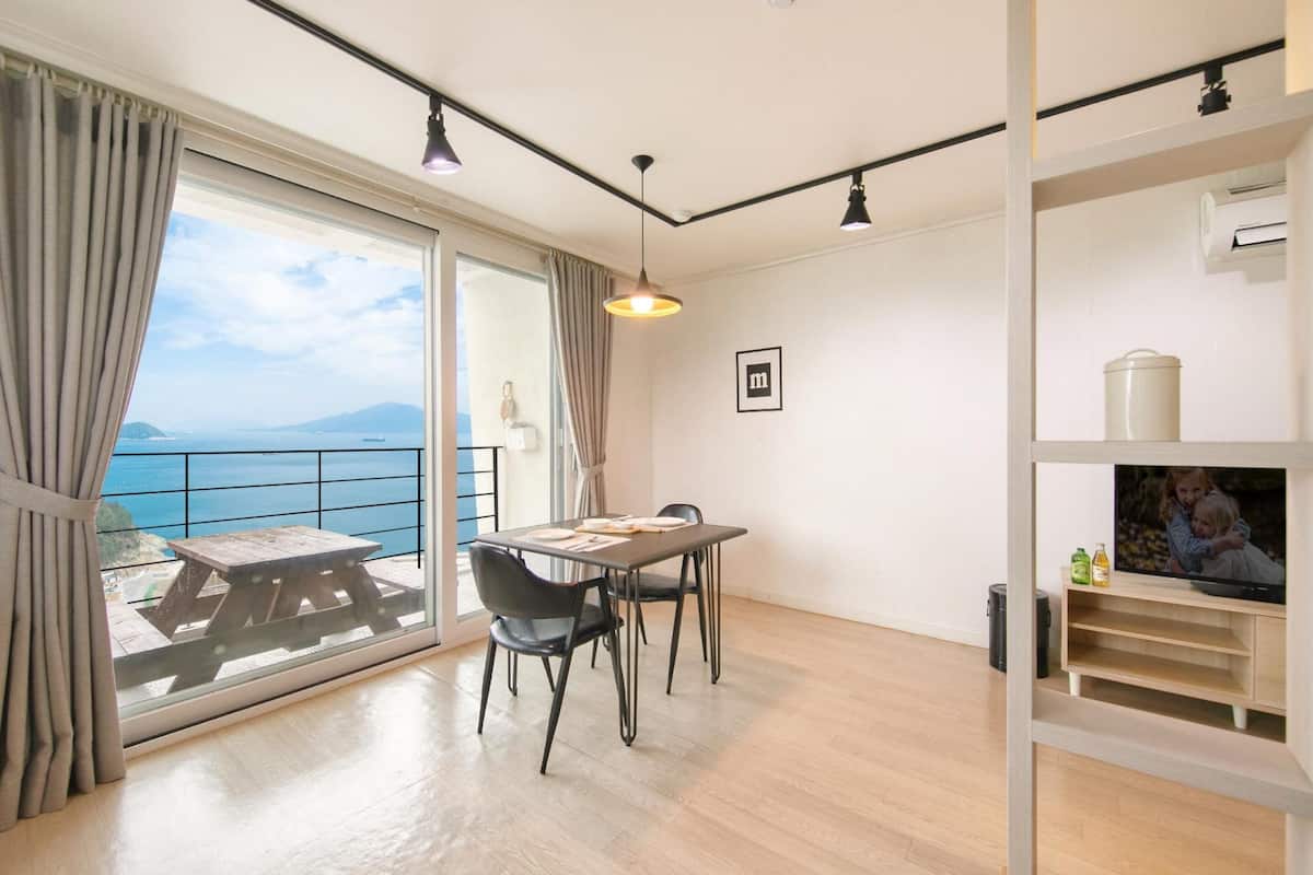 A modern room with a wooden dining table, two chairs, a large window with a view of the sea, and a TV on a shelf.