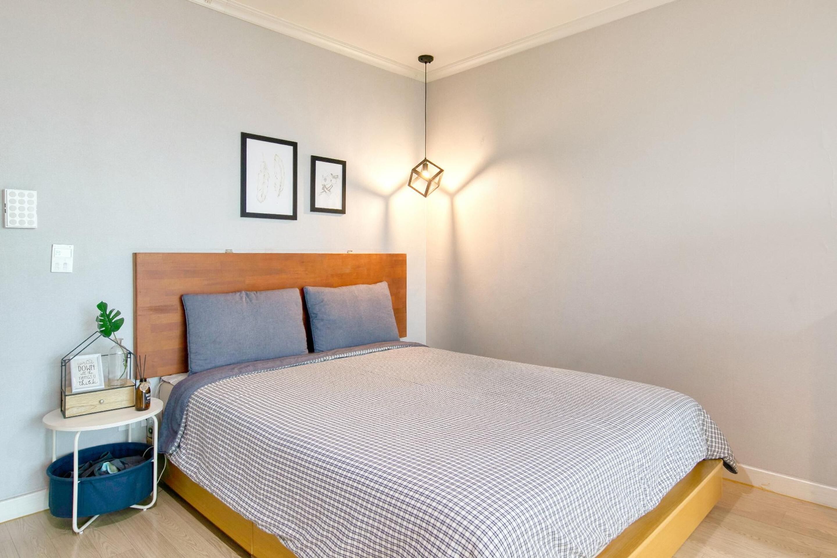 A bedroom with a wooden headboard, a checkered bedspread, a bedside table with a lamp, and a wall-mounted light fixture.