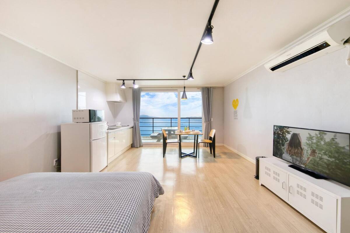 A modern hotel room with a bed, a TV, a kitchenette, a dining area with a table and chairs, and a balcony with a view of the sea.