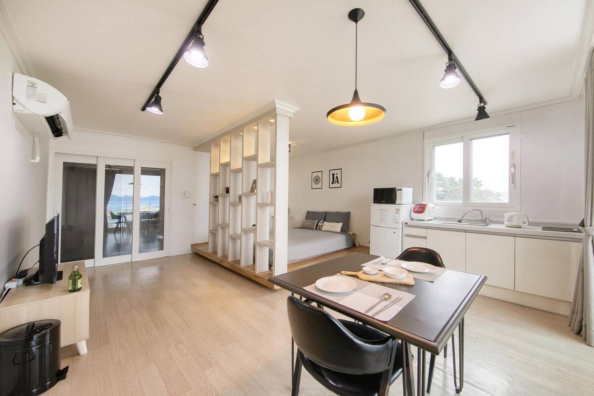 A modern, well-lit room with a dining area, kitchenette, and a sliding glass door leading to a balcony.