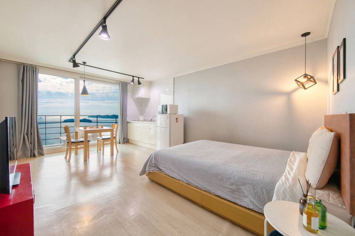 A modern hotel room with a large bed, a dining area with a view, a kitchenette, and a balcony with a view.