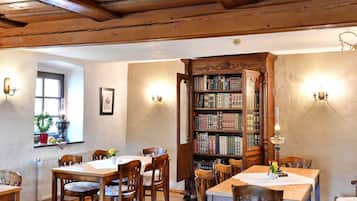 Ein Zimmer mit Holzbalken, einem großen, offenen Bücherregal und einer Essecke mit Tischen und Stühlen.
