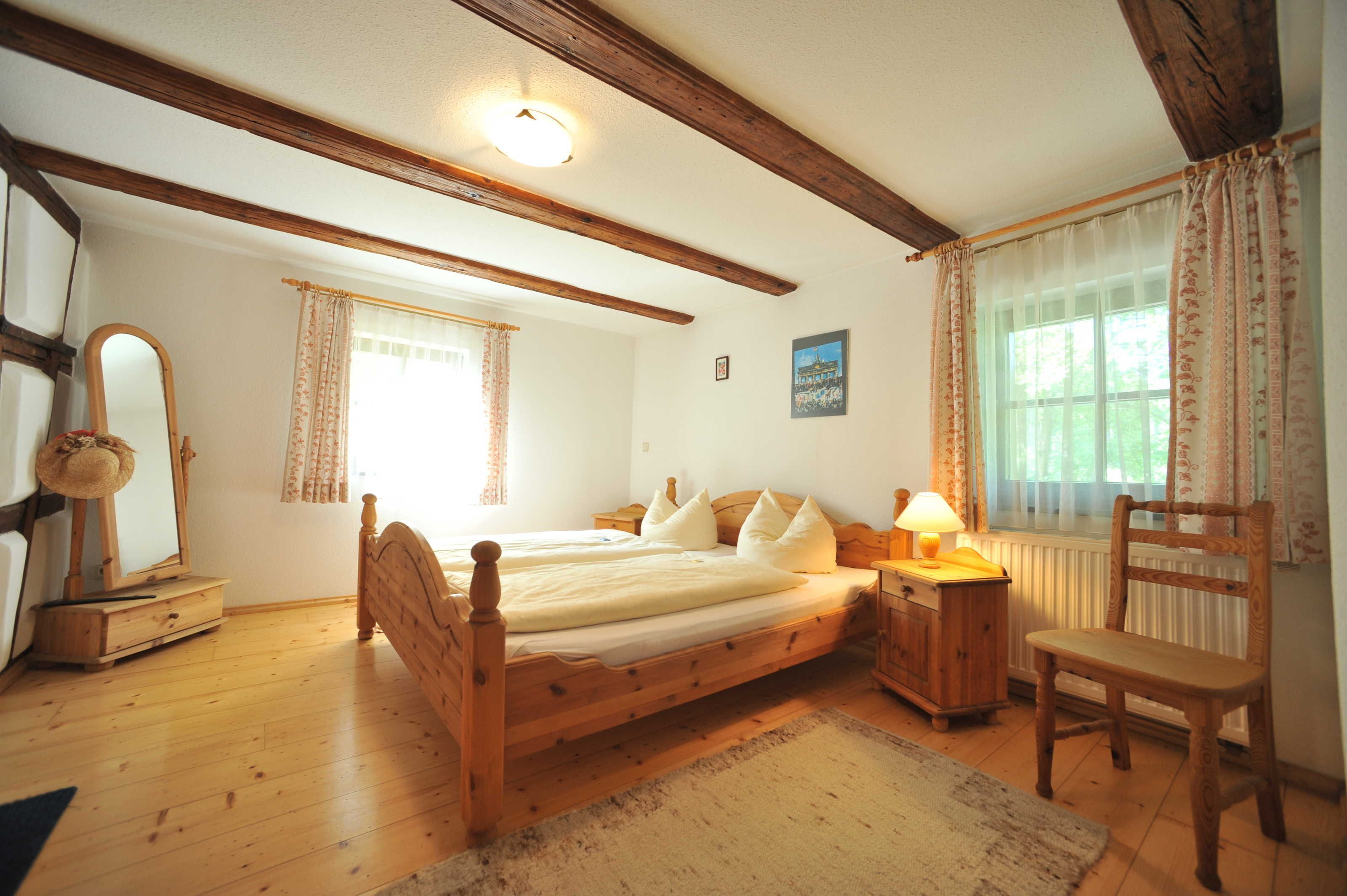 Une chambre à coucher avec un lit en bois, une chaise, une table de chevet, une lampe, un miroir et une fenêtre avec des rideaux.