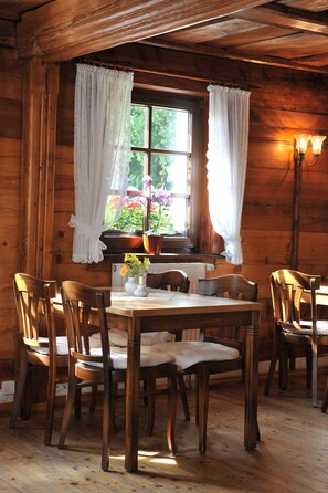 Ein Holzblockhaus-Interieur mit Esstisch, Stühlen, einem Fenster mit Vorhängen und einer Topfpflanze.