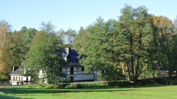 Une maison traditionnelle avec un toit sombre, entourée d’un paysage verdoyant et luxuriant ainsi que d’arbres.