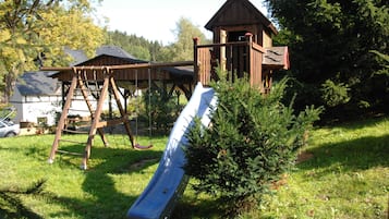 Une aire de jeux avec un toboggan, une structure d’escalade et un petit bâtiment en bois.